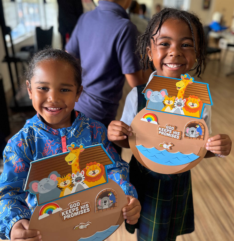 Two smiling young children hold up paper crafts of Noahs Ark decorated with animal faces and rainbows. The crafts say “God Keeps His Promises.” Other people are blurred in the background indoors.
