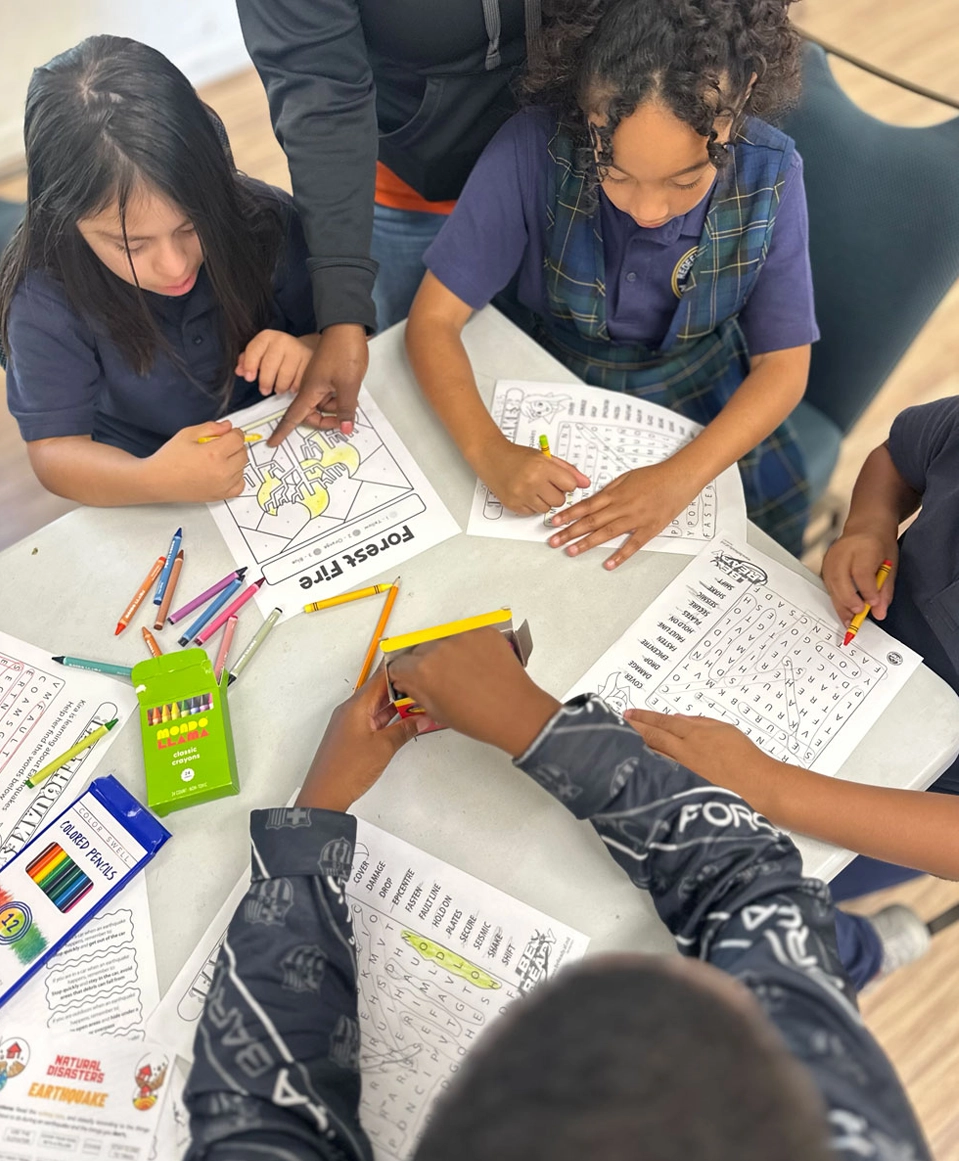Four children sit around a table, coloring and working on word search puzzles with colored pencils. A box of Crayola colored pencils and completed pages are also visible on the table.
